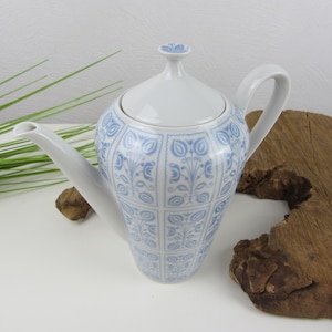 Pode incluir: Bule de cerâmica branca com um padrão floral azul. O bule tem tampa e alça. O design é um padrão repetitivo de flores e folhas estilizadas. O bule está sobre uma superfície branca com um pedaço de madeira.