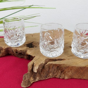 May include: Three clear, cut-glass tumblers with intricate starburst and floral patterns. The glasses are displayed on a natural wood slab against a red background. The glasses are approximately 7.6 cm tall.