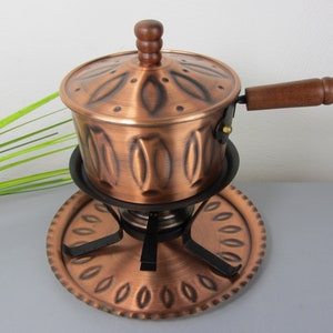 以下が含まれることがあります： 木製のハンドルと装飾的なデザインが施された銅製のフォンデュ鍋。鍋は黒い金属製のスタンドの上に置かれ、銅製のベースがあります。