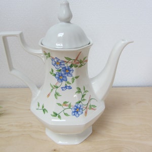 May include: White porcelain coffee pot with a floral design featuring blue flowers and green leaves. The pot has a lid with a knob on top.