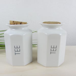 Può includere: Due barattoli di tè in ceramica bianca con coperchi in sughero. Ogni barattolo ha una forma esagonale e la parola "TEE" stampata in nero sul davanti. I barattoli sono su una superficie chiara.