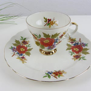 Può includere: Un set vintage di tazza e piattino da tè. La tazza e il piattino in porcellana bianca presentano un motivo floreale con fiori rossi, blu e gialli. La tazza ha una base e un manico color oro, e il piattino ha un bordo dorato. Un classico set da tè.