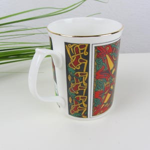 May include: White ceramic mug with a gold rim and a white handle. The mug features a colourful Celtic design in red, yellow, and green on a black background. The mug is a standard size for tea or coffee.