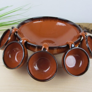 Può includere: Un set di sei tazze in ceramica marrone terracotta con manici, disposte attorno a una ciotola in ceramica marrone terracotta più grande con un bordo nero. Le tazze e la ciotola sono tutte smaltate.