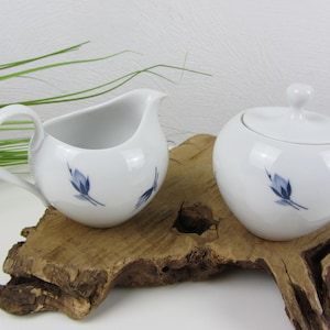 Könnte beinhalten: Ein weißes Sahnegießer- und Zuckergefäß-Set mit blauem Blumendesign. Das Set steht auf einem Holzsockel.