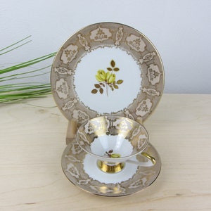 May include: A white teacup and saucer set with a gold trim and a floral design. The saucer has a delicate pattern of gold leaves and flowers. The teacup has a single yellow rose with green leaves.