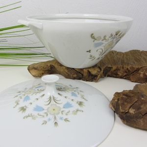 Può includere: Ciotola da portata in ceramica bianca con coperchio, con un motivo floreale blu, beige e verde. La ciotola ha due piccole maniglie e poggia su un pezzo di legno. Il coperchio è davanti alla ciotola.