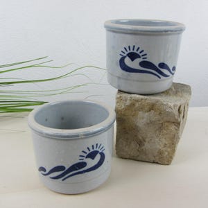 May include: Two small, light blue ceramic pots with a dark blue sun and wave design. The pots have a rounded shape and a slightly flared rim. One pot sits on a stone block, while the other is on a white surface. The pots are likely for decorative or functional use.