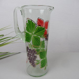 Peut inclure: Une carafe en verre transparent avec une poignée, ornée de feuilles vertes, de fleurs rouges et de grappes de raisin violet. La carafe est haute et élancée, idéale pour servir des boissons. Le design est peint à la main, ajoutant une touche artistique à la verrerie.