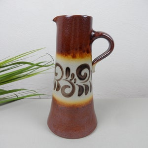 Könnte beinhalten: Eine hohe, braune Keramikkrug mit Henkel und Ausguss. Der Krug hat ein dekoratives Band mit wirbelnden, abstrakten Designs in Braun und Creme. Der Boden ist in einem dunkleren Braunton gehalten, und der Krug ist etwa 25 cm hoch.