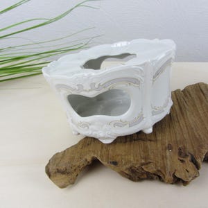 May include: White porcelain tissue box holder with ornate detailing and a decorative design. The holder features a scalloped top and a heart-shaped opening on the front. The design includes grey and gold accents.