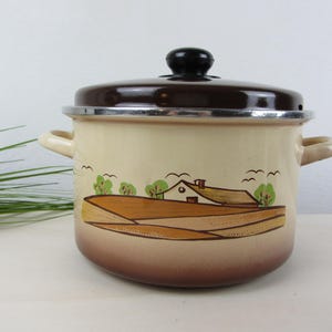 May include: A cream-coloured enamel pot with a dark brown lid and a matching brown gradient base. The exterior features a painted landscape illustration of a house, fields, and trees. It includes two side handles and a black knob on the lid.
