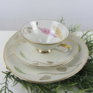 Può includere: Un servizio da tè vintage composto da tazza, piattino e piatto. Il set in porcellana bianca presenta bordi dorati e delicati disegni di piume rosa, verdi e dorate. La tazza ha un manico dorato.