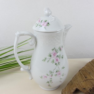 Può includere: Caffettiera in porcellana bianca con coperchio, ornata con motivi floreali rosa e foglie verdi. La caffettiera ha un manico curvo e un beccuccio. Il coperchio è sormontato da un pomello decorativo.