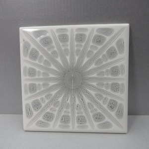 May include: Square white tile with a gray starburst design. The starburst pattern radiates from a central point, with textured lines and shapes. The tile is likely ceramic or porcelain, suitable for home decor or craft projects.