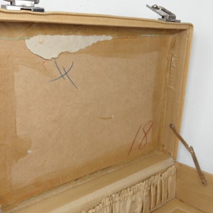 Pu&ograve; includere: Una valigia vintage beige aperta con una fodera in carta marrone. La fodera ha una "X" scritta a mano e il numero "18" in inchiostro rosso.