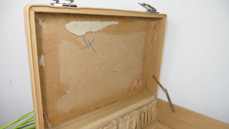 Pu&ograve; includere: Una valigia vintage beige aperta con una fodera in carta marrone. La fodera ha una "X" scritta a mano e il numero "18" in inchiostro rosso.
