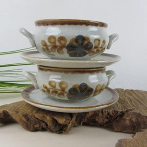 Pode incluir: Duas tigelas de sopa de cerâmica com pires a condizer empilhadas. As tigelas são cinza claro com bordas e alças marrons. Cada tigela apresenta um desenho floral em marrom e cinza escuro. Sobre um pedaço de madeira.