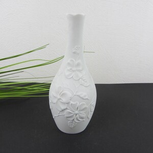 Könnte beinhalten: Eine weiße Porzellanvase mit schlankem Hals und bauchigem Fuß, die mit einem erhabenen Reliefdesign aus Blumen und Blättern verziert ist. Die matte Oberfläche betont die filigranen floralen Texturen, die in den Keramikkörper eingearbeitet sind.