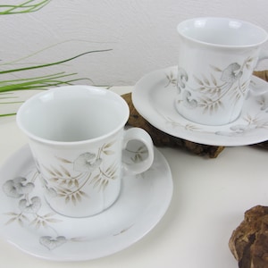May include: Two white porcelain coffee cups and saucers with a floral design. The cups and saucers feature a delicate floral pattern in shades of brown and grey.