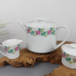 May include: White porcelain teapot, creamer and sugar bowl with a floral design featuring pink roses and green leaves. The set is displayed on a wooden surface.