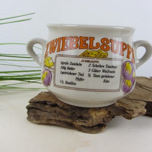 Può includere: Una ciotola per zuppa in ceramica bianca con due manici, caratterizzata dalla scritta ZWIEBELSUPPE in arancione e da una ricetta stampata in nero. La ciotola è decorata con illustrazioni di cipolle, burro e fette di pane tostato.