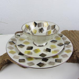 Könnte beinhalten: Ein Vintage-Teeservice, bestehend aus einer Tasse, Untertasse und Teller. Die weißen Porzellanteile weisen ein sich wiederholendes Muster aus schwarzen und gelben Quadraten mit goldenem Rand auf. Die Tasse hat einen Henkel und steht auf der Untertasse.