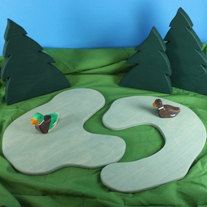 Könnte beinhalten: Holzspielzeug-Set mit zwei Entenfiguren auf hellblauen, teichförmigen Plattformen, mit drei grünen Baum-Ausschnitten im Hintergrund. Die Enten haben braune, grüne und orangefarbene Details. Der Hintergrund ist grün und blau.