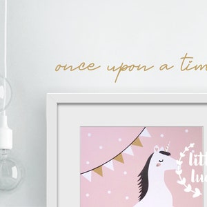 Könnte beinhalten: Ein weißer Rahmen mit rosa Hintergrund, auf dem ein weißes Einhorn mit goldenem Horn und schwarzer Mähne zu sehen ist. Das Einhorn befindet sich unter einer Girlande aus goldenen und weißen Dreiecksflaggen. Der Text "little luck" steht in einem weißen Kreis mit grünem Blattrand. Der Text "once upon a time..." ist in Gold über dem Rahmen geschrieben.