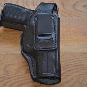Può includere: Fondina in pelle nera con clip in metallo nero, progettata per contenere una pistola. La fondina è mostrata su una superficie di legno. La fondina ha una forma sagomata per adattarsi all'arma da fuoco.