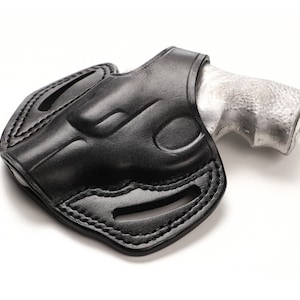 Puede incluir: Funda de cuero negro con una pistola plateada parcialmente insertada. La funda presenta costuras detalladas y trabillas para el cinturón. La imagen está sobre un fondo blanco, destacando el diseño y la artesanía del producto.