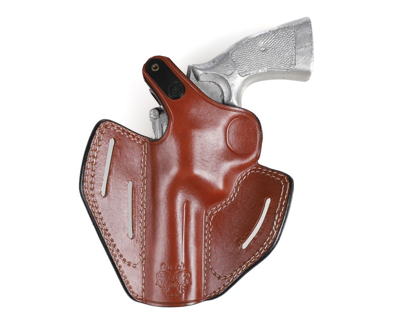 May include: A brown leather holster with a silver handgun. The holster has white stitching and a black border. The handgun is partially inserted into the holster, with the grip visible. The holster is designed to hold a firearm.