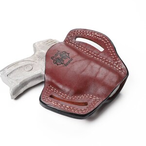 May include: A burgundy leather holster with white stitching and a handle. The holster has the text "Pusch Holster" and a logo. A silver-colored gun is next to the holster.