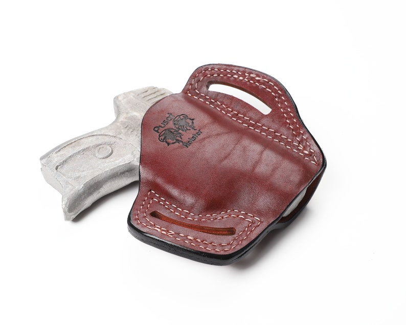 May include: A burgundy leather holster with white stitching and a handle. The holster has the text "Pusch Holster" and a logo. A silver-colored gun is next to the holster.