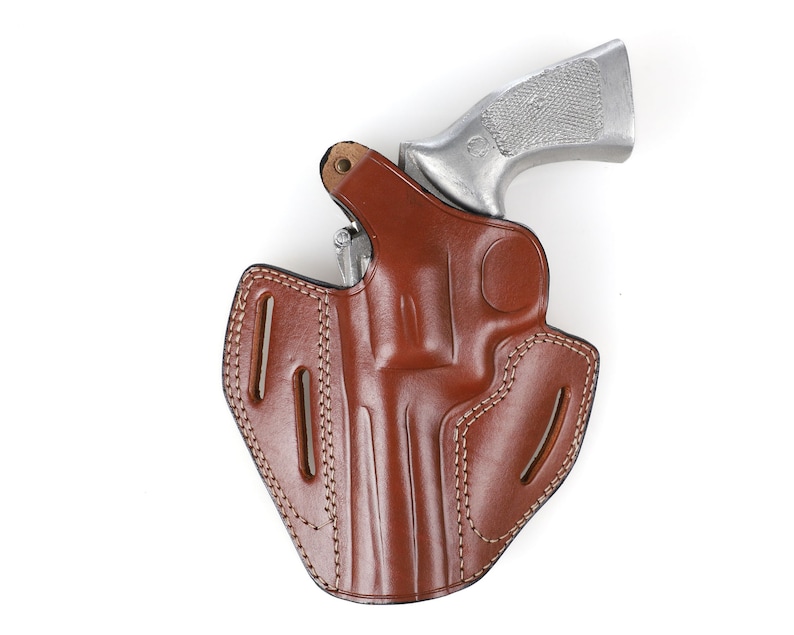 May include: A brown leather holster with a silver handgun. The holster has a molded design with visible stitching and belt loops. The handgun's grip is visible, and the overall image is set against a white background.
