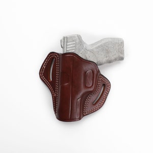 Puede incluir: Una funda de cuero marr&oacute;n con un dise&ntilde;o cosido y recortes para una pistola. La funda est&aacute; vac&iacute;a y la pistola no es visible.