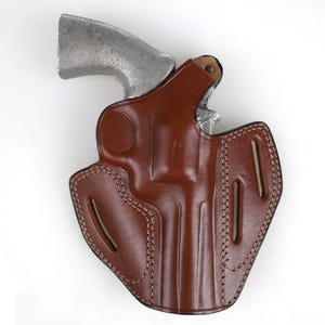 Puede incluir: Una funda de cuero marr&oacute;n con un rev&oacute;lver plateado. La funda tiene costuras decorativas y recortes. El rev&oacute;lver est&aacute; parcialmente insertado, mostrando el ca&ntilde;&oacute;n y el cilindro.