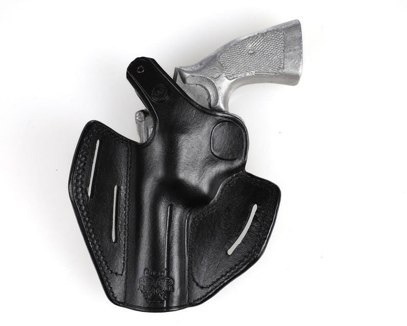 May include: A black leather holster with a silver handgun. The holster has a molded design with stitched edges and vertical slots. The handgun is partially inserted into the holster, with the grip visible.