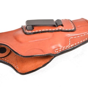 May include: A brown leather holster with white stitching and a black metal clip. The holster is designed to be worn on the belt. The interior is a lighter shade of brown. The holster is made of leather.
