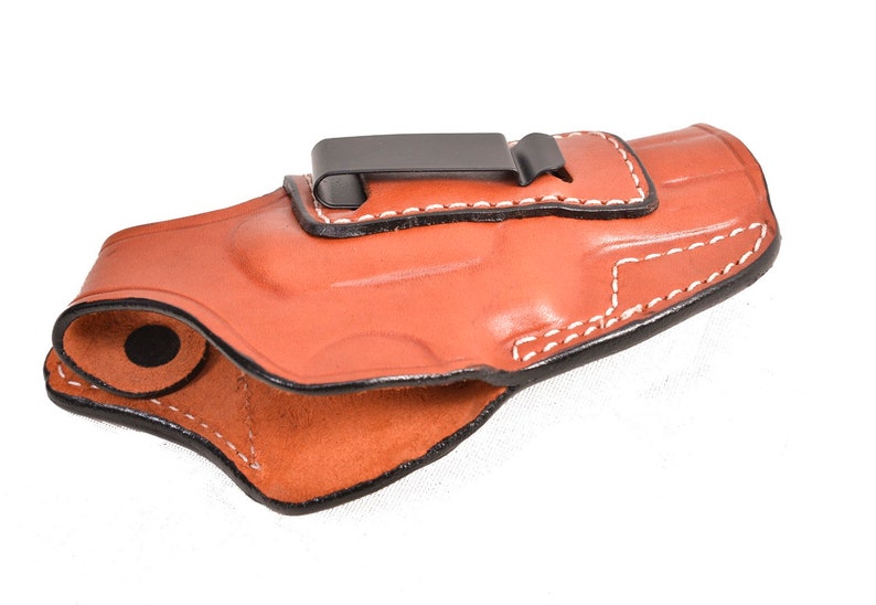 May include: A brown leather holster with white stitching and a black metal clip. The holster is designed to be worn on the belt. The interior is a lighter shade of brown. The holster is made of leather.