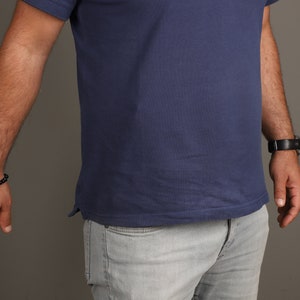 May include: A man wearing a blue short-sleeved polo shirt and light gray jeans. He is wearing a black beaded bracelet on his left wrist and a black watch on his right wrist.