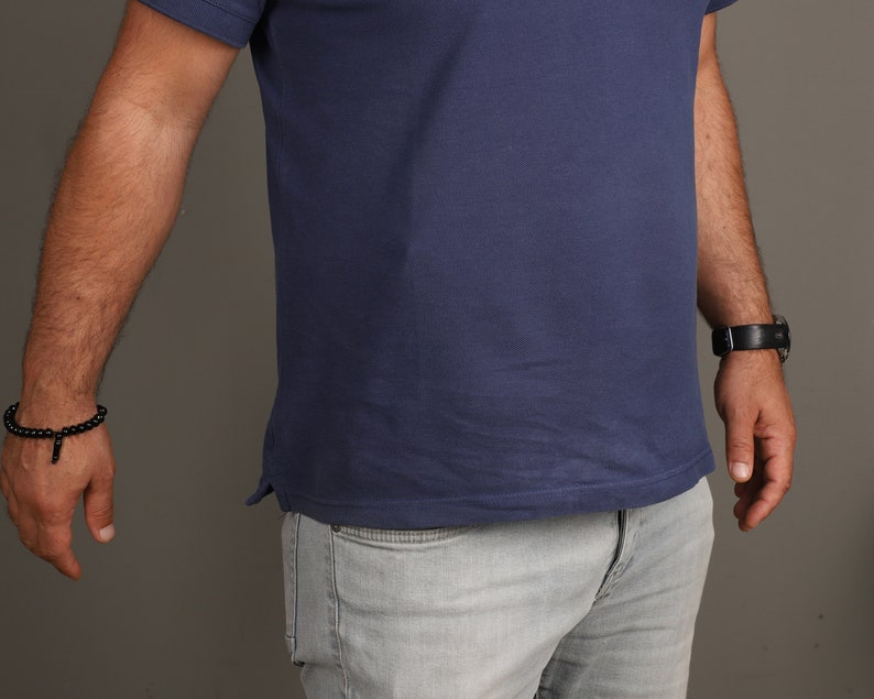 May include: A man wearing a blue short-sleeved polo shirt and light gray jeans. He is wearing a black beaded bracelet on his left wrist and a black watch on his right wrist.