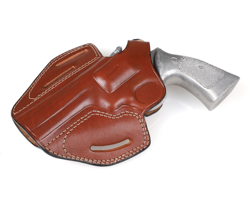 May include: A brown leather holster with white stitching, holding a silver handgun. The holster has a molded design and a belt loop. The handgun's grip is textured.