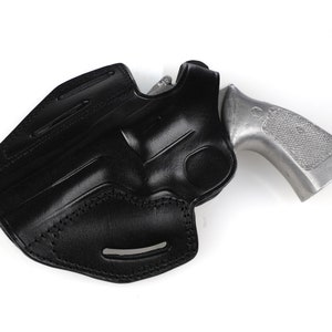 May include: A black leather holster with a silver-colored handgun. The holster is molded to the shape of the gun and features stitching along the edges. The gun's grip is visible, and the overall design suggests a secure and functional carrying accessory.