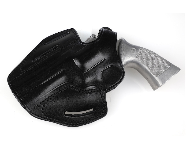 May include: A black leather holster with a silver-colored handgun. The holster is molded to the shape of the gun and features stitching along the edges. The gun's grip is visible, and the overall design suggests a secure and functional carrying accessory.