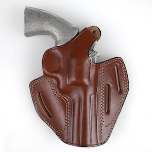 Könnte beinhalten: Ein braunes Lederholster mit Nähten für einen Revolver. Das Holster hat eine Klappe, die den Abzugsschutz bedeckt, und eine Gürtelschlaufe auf der Rückseite.
