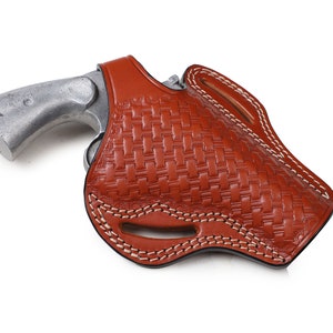 Może przedstawiać: Brązowa skórzana kabura z tkanym wzorem i białymi szwami. Wewnątrz kabury znajduje się srebrny rewolwer.