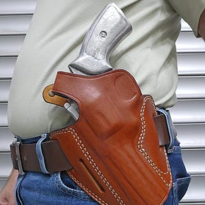 May include: A brown leather holster with white stitching, holding a silver handgun. The holster is attached to a brown belt worn with blue jeans. The handgun's grip is visible above the holster.