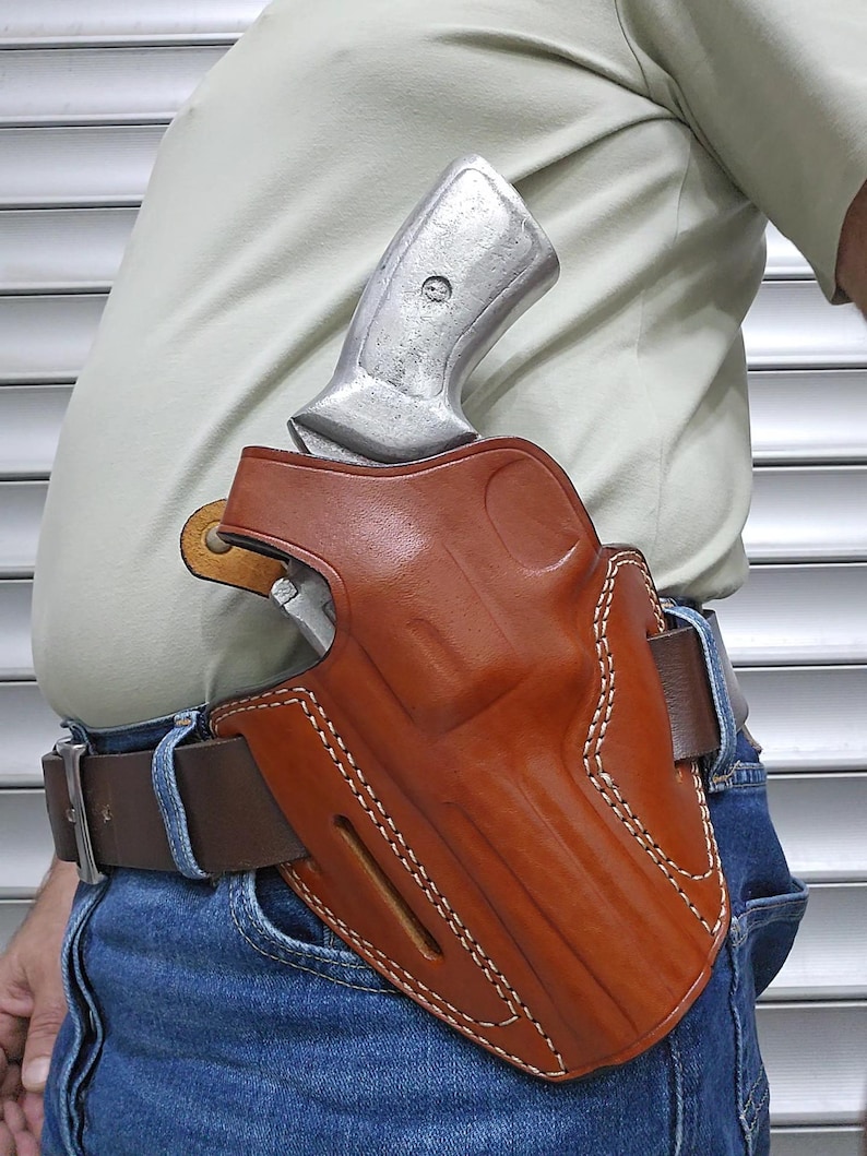 May include: A brown leather holster with white stitching, holding a silver handgun. The holster is attached to a brown belt worn with blue jeans. The handgun's grip is visible above the holster.