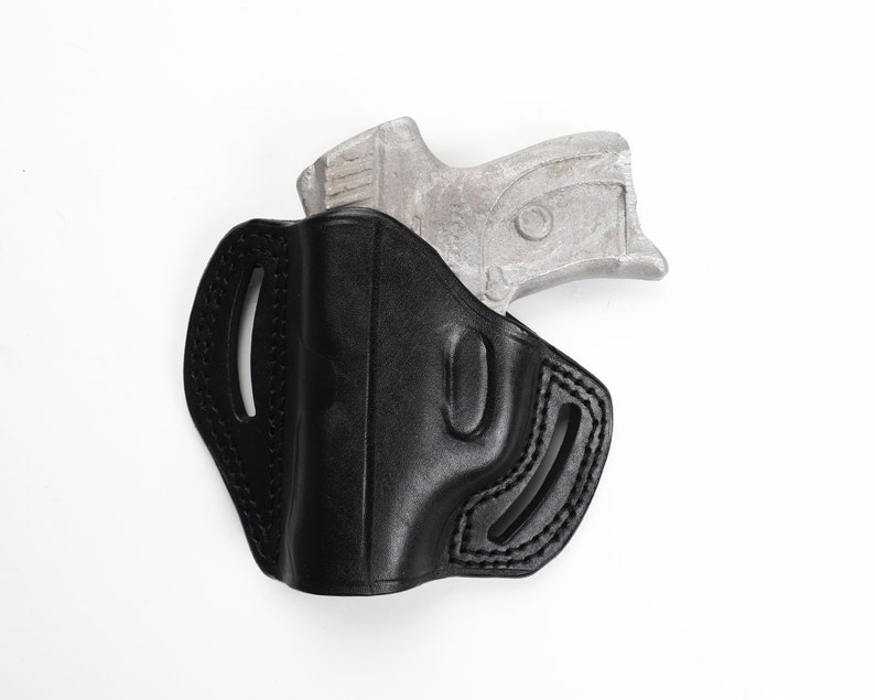 May include: Black leather holster with stitched detailing, designed to hold a handgun. The holster has a molded shape and features cutouts for belt attachment. A silver-colored handgun is partially visible.